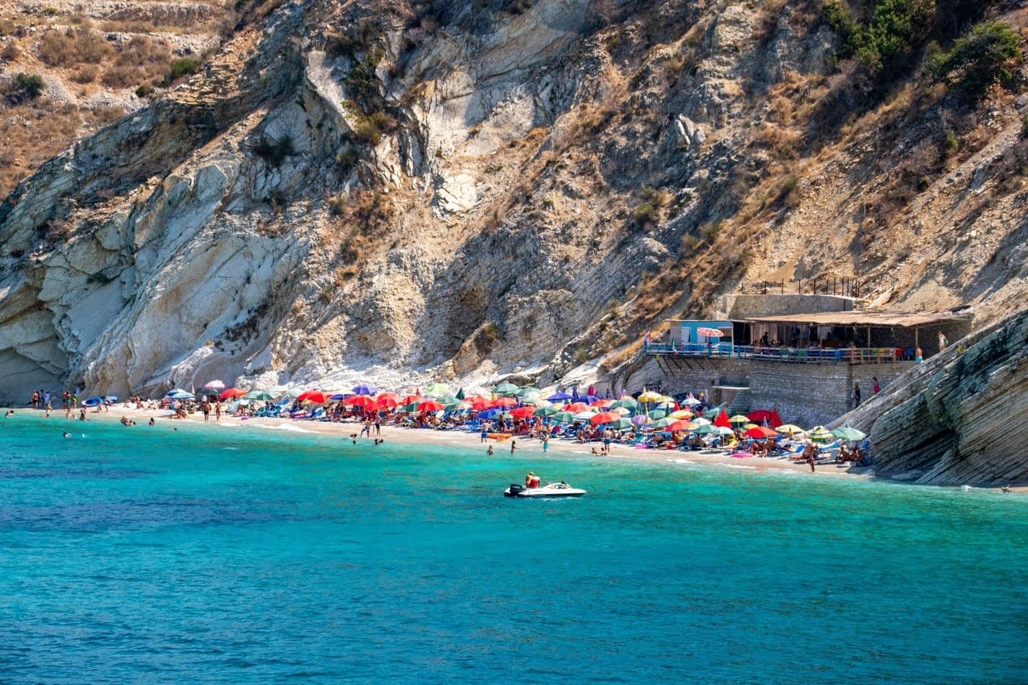 Shpella e Pëllumbave — a cliff-backed pebble cove with turquoise water and a beach bar built into the rock