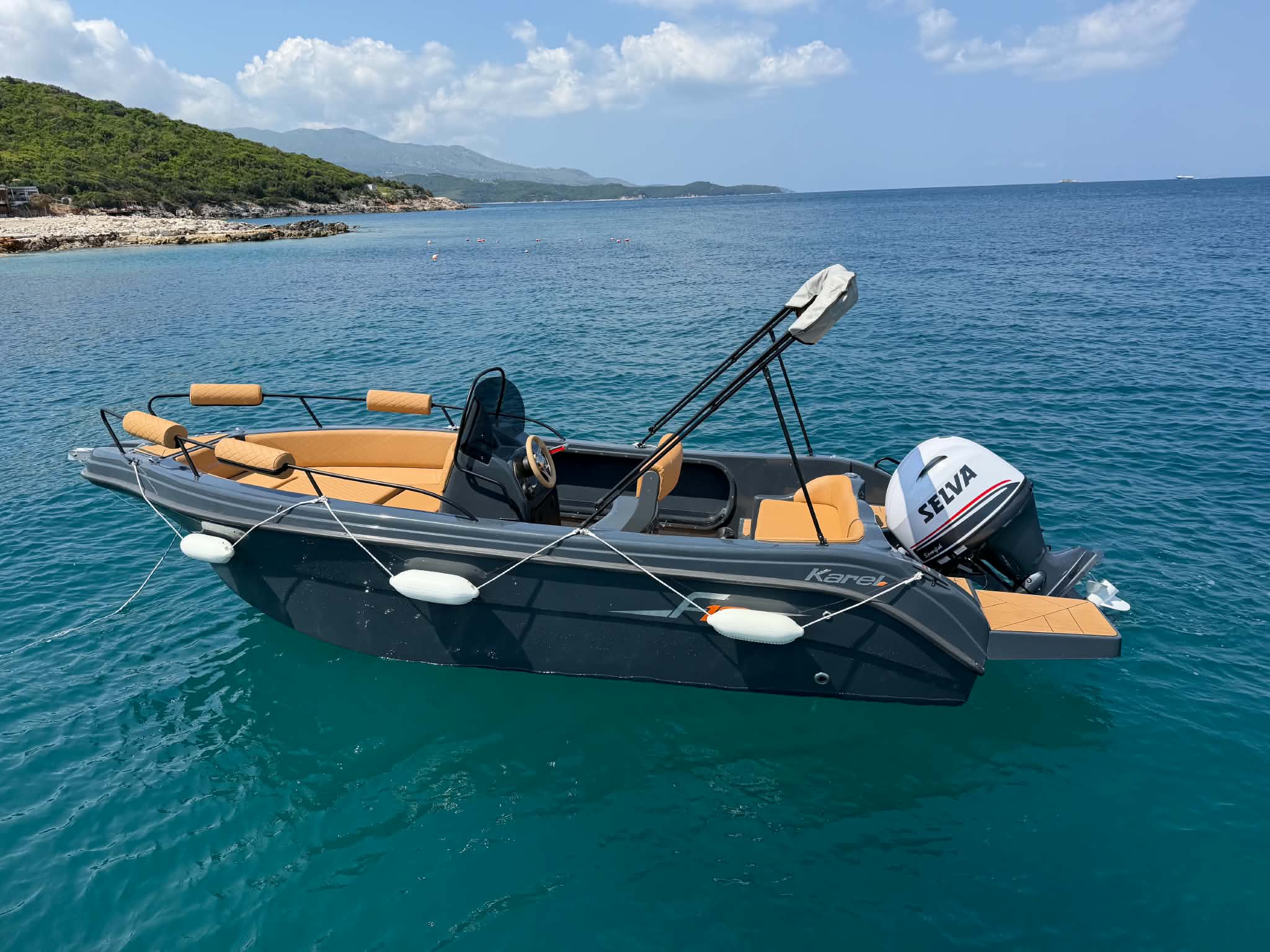 Charcoal-grey open boat with tan cushions and a Selva outboard, anchored over deep teal water below the pine-covered Ionian coast