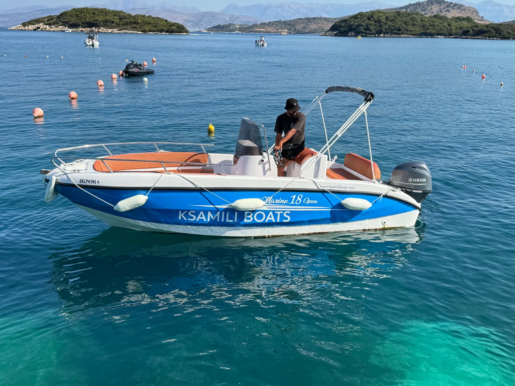 Skipper standing at the console of a Marina 18 Open in Ksamili Boats livery, anchored over clear turquoise water with the Ksamil islets behind